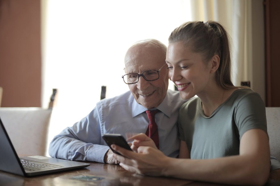 jeune-femme-positive-montrant-des-photos-sur-smartphone-a-senior-homme-assis-a-l-ordinateur-portable